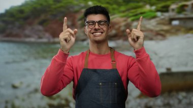 Gözlüklü ve önlüklü genç adam sahilde neşeyle gülümsüyor manzaralı bir arka planda.