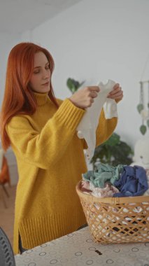 Evde rahat bir çamaşırhanede elbiselerini katlayan, kızıl saçlarını sergileyen ve sarı bir kazak giyen, yeni temizlenmiş çamaşır sepetleriyle çevrili bir kadın..