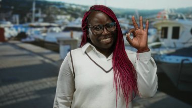 Arka planda tekneleri ve deniziyle bir limanda pembe örgülü gülümseyen kadın, açık deniz ortamında mutluluk ve tarz yayıyor..