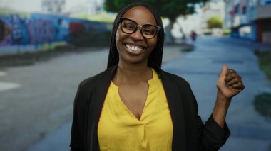 Şehir merkezinde gülümseyen gözlüklü ve sarı bluzlu bir kadın modern yaşam tarzını tasvir eden canlı bir şehir manzarasında açık hava mutluluğunu sergiliyor.