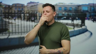 Şehir dışında hapşıran genç bir adam yeşil gömlekli ve dövmeli bir adam. Gün boyunca bulanık şehir arka planına karşı görünür..