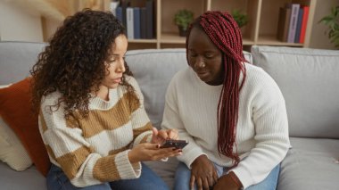 Two women sitting on a cozy sofa in a modern living room engaged in conversation, sharing an intimate moment as they enjoy their friendship and discuss using a smartphone.