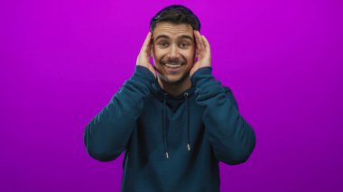 Deniz mavisi kapüşonlu genç adam, gülümsüyor ve mor arka planda yüzünü örtüyor, neşe ve neşe ifade ediyor, ellerini yüzünü çerçeveliyor..