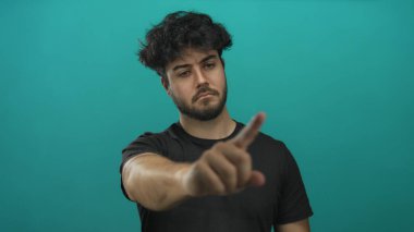 Sakallı genç adam mavi arka plana karşı küçümseyici bir jest yapıyor. Ciddi görünüyor ve odaklanmış. Hareketi, karar ya da reddedilmeyi ima ediyor.