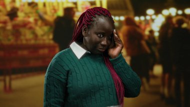 Kırmızı örgülü, yeşil kazaklı bir kadın, açık hava panayırında duruyor, bulanık ışıklar ve insanlarla çevrili, konuşurken şaşırmış görünüyor..