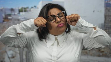 Gözlüklü genç bir kadın, bir inşaat alanında ağlama hareketi yapıyor, dışarıda duygularını ifade ediyor, Latin, artı beden, beyaz gömlek giyiyor..