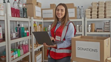 İç mekândaki hayır kurumlarında dizüstü bilgisayar kullanan genç bir kadın. Etrafı kutularla ve malzeme raflarıyla çevrili. Kırmızı yelek giymiş, insani yardım organizasyonları düzenliyor.