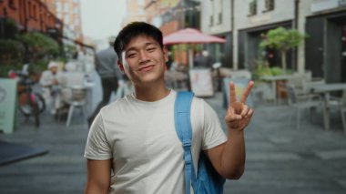 Genç adam gülümsüyor ve açık bir kahve dükkanının terasında barış işareti yapıyor. Kalabalık bir şehir restoranında rahat bir atmosferi var..