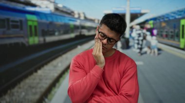 Kırmızı kazaklı genç adam hareketli bir tren istasyonunda düşünceli görünüyor. Şehir içi banliyö seansının arka planında bulanık bir aktivite olduğunu gösteriyor..