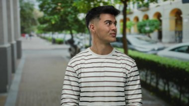 Çizgili tişörtlü genç bir adam şehir caddesinde gülümsüyor, yeşilliklerle çevrili, dışarıda çekici bir şehir portresi sunuyor..