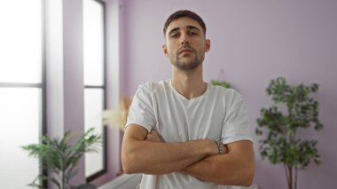 Yakışıklı, İspanyol asıllı bir adam, kolları kapalı, şık bir apartman dairesinde, etrafı bitkilerle ve modern dekorlarla çevrili bir şekilde duruyor..