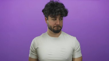 Beyaz tişörtlü, sakallı, genç İspanyol adam canlı mor arka plana karşı duruyor. Düşünceli bir yüz ifadesi var. Modern gündelik stil ve izolasyon kavramı sergiliyor.