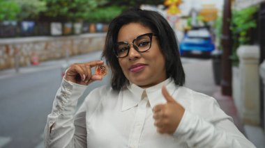 Şehir sokağında elinde kripto para tutan genç bir kadın kendine güvenen bir ifadeyle açık bir şehirde kripto şevki ve modern finansmanı vurguluyor..