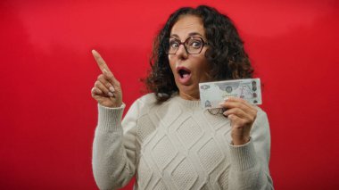 Orta yaşlı bir kadın elinde Uae Dirham banknotu, şaşırmış yüz ifadesi, kırmızı arka plan, gözlük ve süveter giyiyor,.