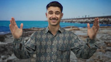 Genç İspanyol adam açık mavi bir deniz ve gökyüzü ile güneşli bir sahil tarafında, açık bir el hareketi yapıyor, desenli bir gömlek giyiyor, neşeli ve rahat bir açık hava atmosferi taşıyor..