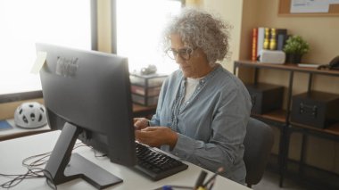 Modern ofiste akıllı telefon kullanan, gri saçlı bir kadın bilgisayarlı, kitaplı ve kasklı bir masada otururken selfie çekiyor, gülümsüyor ve içeride çalışıyor..