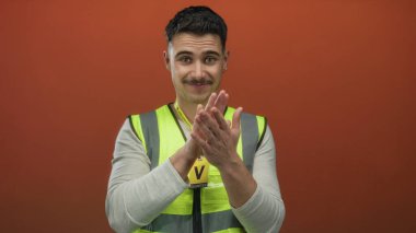 Yansıtıcı yelek giyen genç İspanyol adam izole edilmiş turuncu arka plana karşı gönüllü olarak işaret ediyor ve dost ifadesiyle alkışlıyor..