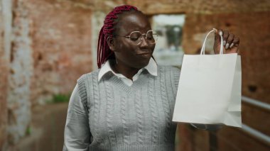 Bir inşaat alanında gülümseyen ve beyaz bir alışveriş torbasını inceleyen bir kadın, kırsal tuğla bir arka planda dururken gözlüğünü ve gri kazağını sergiliyor..