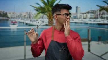 Deniz kıyısında bir limanda elinde makasla dolaşan gözlüklü ve önlüklü genç adam bir tekne ve palmiye ağacına karşı neşeli bir ifade sergiliyor..