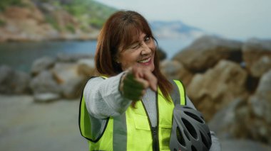 İspanyol kıdemli bir kadın plajda canlı bir şekilde kameraya göz kırpıyor ve onu işaret ediyor, yüksek görüş mesafesi olan bir yelek giyiyor ve kayalıkların yanında bisiklet kaskı tutuyor..