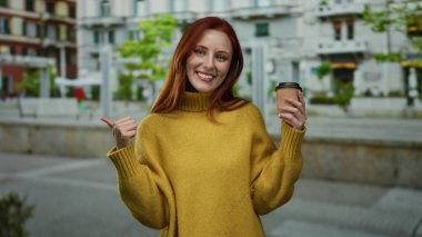 Şehir caddesinde kahveyle gülümseyen, sarı kazak giyen, baş parmak kaldıran, kızıl saçlı, ağaçlı ve mimarlı şehir manzaralı bir kadın..