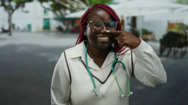 Sokakta steteskoplu gülümseyen kadın gözlüklü ve örgülü saçlarıyla burnuna bakıyor. Arkadaşça ve cana yakın bir tavır sergiliyor..