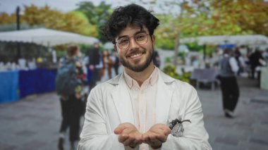 Sakallı ve gözlüklü genç adam hareketli bir sokak pazarında gülümseyen ellerini uzatıyor. Etrafı renkli tezgahlar ve arka plandaki insanlarla çevrili..