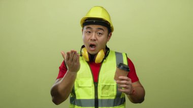 Yorgun Asyalı inşaat işçisi sarı arka planda kahve tutarak esniyor güvenlik ekipmanları ve miğfer takıyor iş sırasında yorgunluk hissi veriyor..