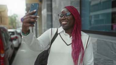 Pembe örgülü bir kadın arka planda arabaları olan bir caddede telefon görüşmesi yapıyor, akıllı telefon kullanıyor ve gülümsüyor..