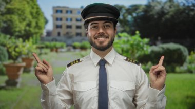 Pilot üniforması ve şapkası içinde sakallı, gülümseyen genç İspanyol adam yeşil bir parkta duruyor. Arka planda bulanık bir bina var..