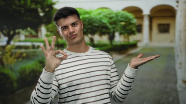 Üniversite bahçesinde jest yapan çizgili tişörtlü genç bir adam akademik ortamda olumlu ifade sergiliyor.