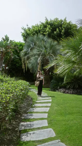 Parlak tropik tatil köyü bahçesinde, güneşli lüks otel ortamında canlı palmiyeler ve egzotik bitkilerle, zarif bir tarzı ve sakin bir atmosferi olan taştan yolda yürüyen bir kadın.