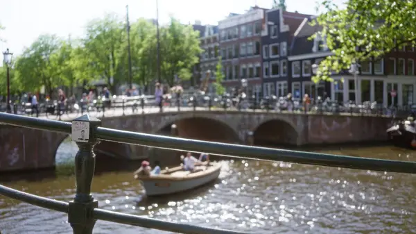 Güneşli bir günde yeşil ağaçların ve geleneksel Hollandalı mimarinin arkaplanına odaklanmış bir tekne ve insanların olduğu bulanık bir kanal manzarası.