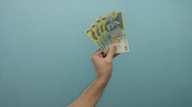 Romen lei banknotlarını mavi arka planda tutan adam, para ve finans gösterisini basit ve izole bir ortamda yapıyor..