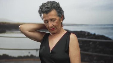 Uzakta sakin bir sahil kenarında boynuna dokunan gri saçlı son sınıf öğrencisi kadın..