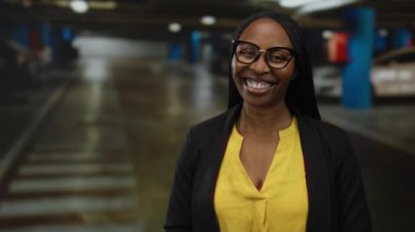 Kapalı otoparkta gülümseyen, sarı tişörtlü ve gözlüklü bir kadın. Arka planda bulanık arabalarla kendinden emin ve neşeli bir tavır sergiliyor..