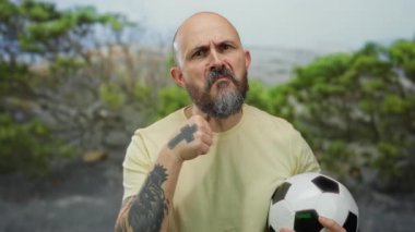 Sakallı kel bir adam parkta futbol topu tutarak tehdit edici hareketlerde bulunuyor. Dışarıdaki duygulara ve dövmelere vurgu yapıyor..