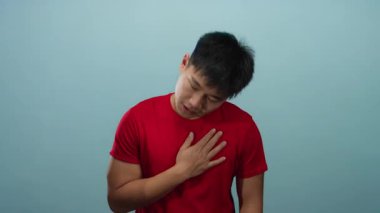 Kırmızı tişörtlü genç adam, elini göğsüne koyup mavi arka plan duvarına yaslanmış düşünceli ve içe dönük görünüyor..