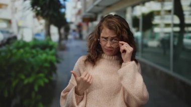 Gözlüklü kadın şehir merkezinde dikilirken açık şehir ortamında ifadesini sorgulayan süveter giyiyor.