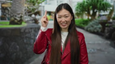 Kırmızı ceketli kadın gündüz vakti şehir sokaklarında gülümsüyor, arkasında ağaçlar ve binalar olan neşeli bir yüz ifadesi sergiliyor..