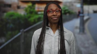 Gözlüklü bir kadın şehir caddesinde çizgili tişört giyer ve arka planda yeşillikle doğal gün ışığında ifadesini göstererek halk önünde konuşma yapmayı önerir.