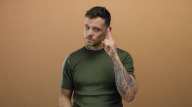 Dövmeli genç İspanyol bir adam, yeşil tişört giyiyor, düşünceli bir şekilde el kol hareketi yapıyor, kahverengi bir duvar arkasında bize dolar tutuyor, izole edilmiş ve mali tefekkür öneriyor..