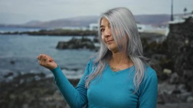 Sahilde başparmağıyla işaret eden bir kadın kayalık deniz manzarasının yanında gri saçlı mavi gömlekli bir kadın.