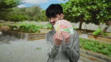 Parlak bir park ortamında Norveçli banknotları elinde tutan adam yemyeşil, parlak doğal bir ortamda gösteriş yapan paralarla güvenle gülüyor..