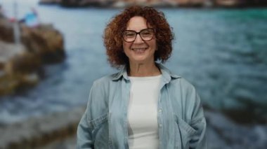 Hispanic woman with curly hair sends a kiss to the camera at a seaside location with rocky shores and blue ocean waves in the background.