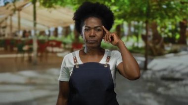 Önlük giymiş bir kadın, şehir caddesinde bulanık ağaçlarla ve açık şehir ortamındaki insanlarla dikilirken kafaya dokunuyor. Düşünceli bir ifade sergiliyor..