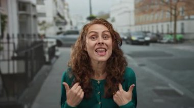 Yeşil kazaklı, kıvırcık saçlı genç bir kadın şehir merkezinde canlı jestler ve yüz ifadeleri sergiliyor..