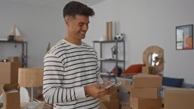 Çizgili kazaklı genç adam oturma odasında gülümsüyor ve dolar sayıyor. Etrafı kutularla çevrili. Yeni ev heyecanını ve geçişi yansıtıyor..