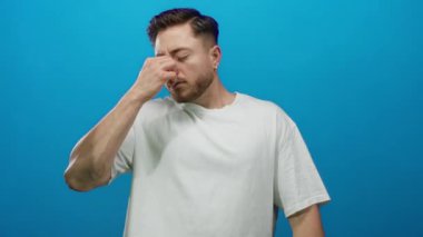 Sakallı genç adam yorgun görünüyor, burnu mavi bir arka plana sürtünüyor, izole edilmiş, günlük beyaz gömlek giyiyor, yorgunluk veya stres taşıyor..