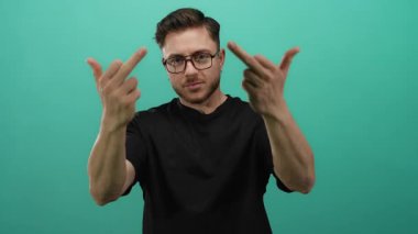 Sakallı genç adam, siyah gömlek ve gözlük takarak yeşil arka planda duran orta parmaklarıyla saldırgan hareketler yapıyor.